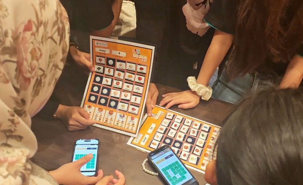 Colleagues gathered around an AR Rojak game board, scanning tiles with their phones